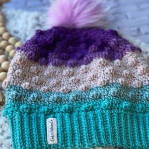 May include: A multi-colored crocheted beanie with a purple, beige, and teal color gradient. The beanie has a fluffy pink pom pom on top and a white label that says "Dukes & Autumn Co."