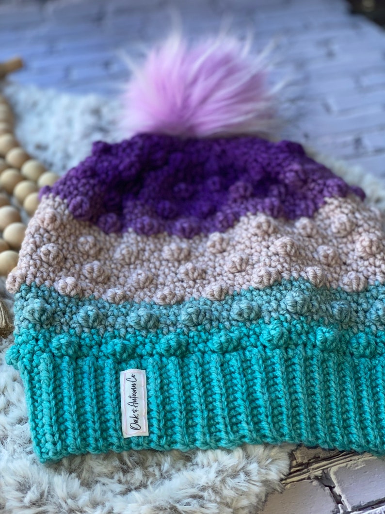 May include: A multi-colored crocheted beanie with a purple, beige, and teal color gradient. The beanie has a fluffy pink pom pom on top and a white label that says "Dukes & Autumn Co."