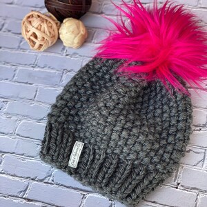 May include: Gray knitted beanie with a large, bright pink faux fur pom pom. The beanie has a small white label that reads "Cider & Hawthorn Co."