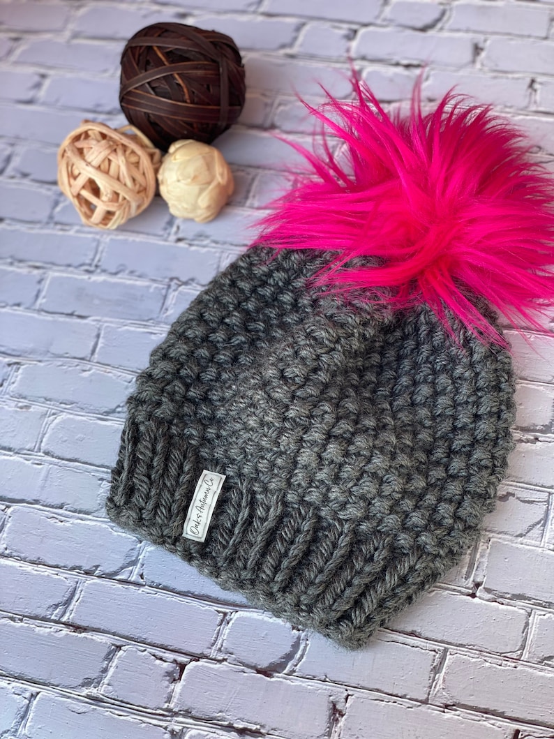 May include: Gray knitted beanie with a large, bright pink faux fur pom pom. The beanie has a small white label that reads "Cider & Hawthorn Co."
