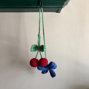 Puede incluir: Un adorno colgante de ganchillo que presenta dos pequeñas esferas rojas que parecen cerezas, unidas a un tallo verde claro con una hoja verde claro. Un lazo de ganchillo azul está unido al tallo, colgando debajo de las esferas rojas.