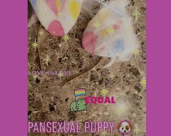 May include: White, fluffy puppy ears with a gold headband. The ears are decorated with pastel colors and a heart. Text on the image includes "LOVE IS LOVE", "EQUAL", and "PANSEXUAL PUPPY EARS".