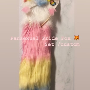 May include: A pansexual pride fox tail and headband set. The tail is pink, yellow, and blue, and the headband has a white fur pom-pom and a pansexual pride flag.
