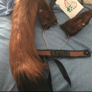 May include: Brown faux fur tail with a black ribbon, a brown faux fur headband with ears, and a brown and black ribbon with a black flower.