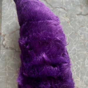 Peut inclure: Un accessoire en forme de queue d'animal, effilé et recouvert d'une longue fausse fourrure violette vive. L'objet présente une pointe incurvée et une texture douce et épaisse, avec une petite attache métallique visible à la base.