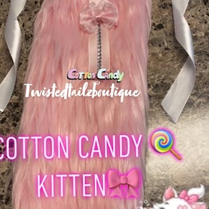 Może przedstawiać: Różowy puszysty ogon z białą wstążką i różową kokardką. Ogon jest oznaczony jako "Cotton Candy Kitten" z różową kokardką i grafiką lizaków.