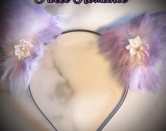 May include: A headband with fluffy, lavender and pink cat ears. Each ear is adorned with a small, white flower with a rhinestone center. The text "Sweet Romance" is written in a cursive font above the ears.