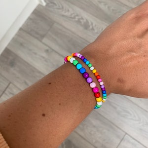 May include: Two colourful beaded bracelets on a wrist. The bracelets are made of small, round beads in a rainbow of colours, including red, orange, yellow, green, blue, indigo, and violet.