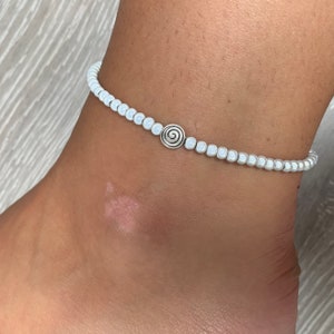以下が含まれることがあります： シルバーの渦巻きチャームが付いた白いビーズのアンクレット。アンクレットは、小さな丸い白いビーズを繋ぎ合わせて作られています。チャームはシルバーの渦巻きデザインです。
