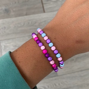 Könnte beinhalten: Zwei Armbänder mit Perlen in einem Farbverlauf von rosa, lila und hellblau.