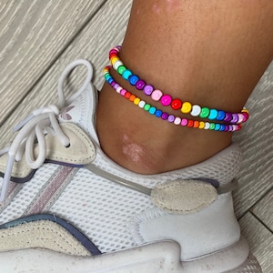 May include: Two colorful beaded anklets on a person's ankle. The anklets are made of small, round beads in a rainbow of colors. The anklets are layered on top of each other, with the top anklet being slightly smaller than the bottom anklet.