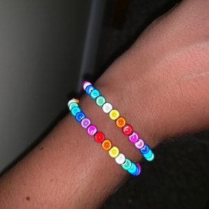Op de afbeelding: Twee regenboogkleurige kralenarmbanden om de pols van een persoon. De armbanden zijn gemaakt van kleine, ronde kralen in verschillende kleuren, waaronder rood, oranje, geel, groen, blauw en paars.