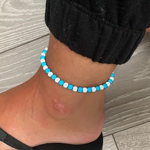 Puede incluir: Un brazalete de tobillo de cuentas blancas y turquesas. El brazalete está hecho de pequeñas cuentas redondas que están enhebradas en un cordón delgado.