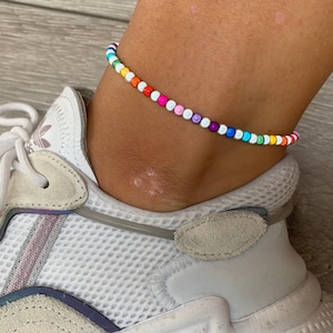 May include: A rainbow beaded anklet with white beads. The anklet is on a person's ankle, and the person is wearing white sneakers.