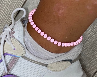 May include: A pink beaded anklet with small, round beads. The anklet is worn on a person's ankle.