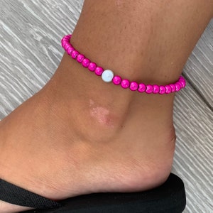 Könnte beinhalten: Ein leuchtend pinkfarbenes Fußkettchen mit einer einzelnen weißen Perle. Das Fußkettchen wird an einem Knöchel getragen, mit einer schwarzen Flip-Flop sichtbar. Die Perlen haben einen Durchmesser von etwa 0,6 cm.