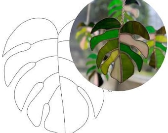 May include: A stained glass window hanging features a green and pink monstera leaf. The leaf is detailed with veins and has a light green and white background.