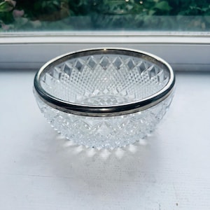 Op de afbeelding: Een heldere glazen kom met een gedetailleerd diamantgeslepen patroon over het buitenoppervlak. De ronde rand van de kom is afgewerkt met een gladde, gepolijste zilverkleurige metalen band die de bovenkant van de glazen kom omsluit.