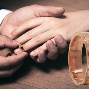 May include: A close-up of a gold wedding band being placed on a finger. The band is simple and elegant, with a smooth finish.
