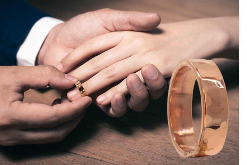 May include: A close-up of a gold wedding band being placed on a finger. The band is simple and elegant, with a smooth finish.