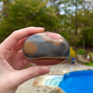 May include: A smooth, round, multi-colored stone with brown, red, and gray bands. The stone is being held in a hand.