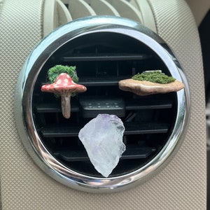 May include: Three car air freshener charms hanging from a car air vent. The charms are a red and white mushroom, a small rock with green moss, and a clear crystal.