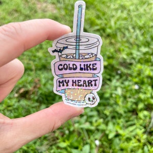 Pu&ograve; includere: Un adesivo olografico di una tazza di caff&egrave; con una cannuccia e un pipistrello che vola sopra. La tazza ha uno striscione rosa che dice "Cold Like My Heart" e un teschio in fondo.