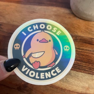 May include: A round sticker with a holographic background featuring a yellow cartoon duck holding a knife. The sticker has the text "I CHOOSE VIOLENCE" in a white circle with black outlines and two skull icons.