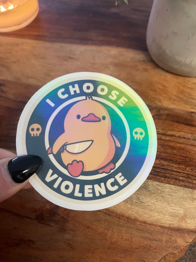 May include: A round sticker with a holographic background featuring a yellow cartoon duck holding a knife. The sticker has the text "I CHOOSE VIOLENCE" in a white circle with black outlines and two skull icons.