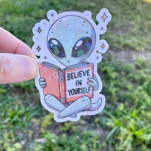 K&ouml;nnte beinhalten: Ein grauer Alien-Cartoon-Charakter liest ein Buch mit der Aufschrift "Glaube an dich selbst". Das Alien sitzt mit gekreuzten Beinen und hat gro&szlig;e Augen und einen kleinen Mund. Das Buch ist rosa und hat ein Herz auf dem Cover.