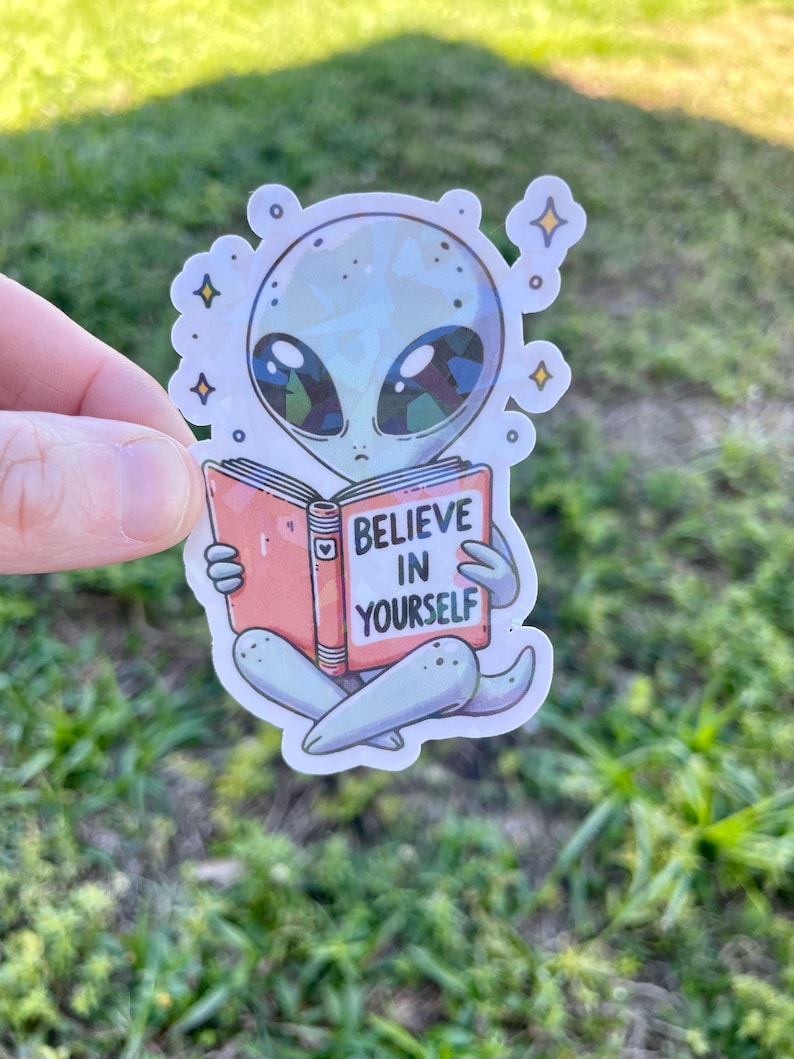 K&ouml;nnte beinhalten: Ein grauer Alien-Cartoon-Charakter liest ein Buch mit der Aufschrift "Glaube an dich selbst". Das Alien sitzt mit gekreuzten Beinen und hat gro&szlig;e Augen und einen kleinen Mund. Das Buch ist rosa und hat ein Herz auf dem Cover.