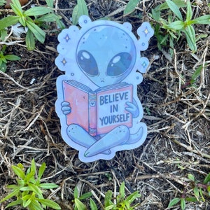 K&ouml;nnte beinhalten: Ein Aufkleber mit einem grauen Au&szlig;erirdischen, der ein Buch liest, auf dem "Glaube an dich selbst" steht. Der Au&szlig;erirdische sitzt mit gekreuzten Beinen und hat einen nachdenklichen Gesichtsausdruck. Der Aufkleber hat eine holografische Oberfl&auml;che und ist von kleinen Sternen umgeben.