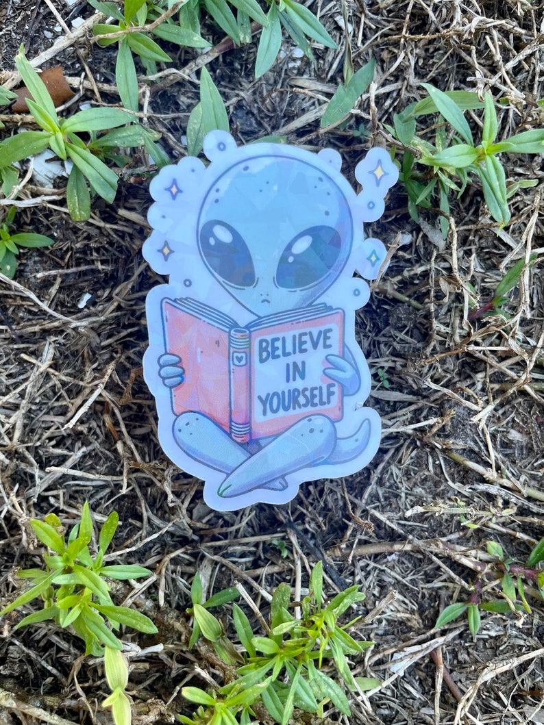 K&ouml;nnte beinhalten: Ein Aufkleber mit einem grauen Au&szlig;erirdischen, der ein Buch liest, auf dem "Glaube an dich selbst" steht. Der Au&szlig;erirdische sitzt mit gekreuzten Beinen und hat einen nachdenklichen Gesichtsausdruck. Der Aufkleber hat eine holografische Oberfl&auml;che und ist von kleinen Sternen umgeben.