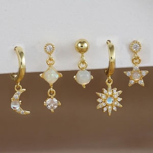 May include: Set of five gold-tone earrings with dangling charms. The charms include a crescent moon, a star, a square, a round opal, and a starburst. Each charm is adorned with clear cubic zirconia stones.