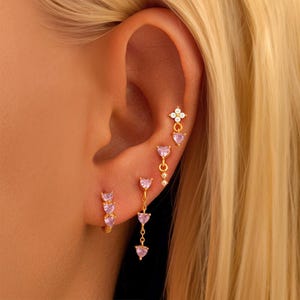 Może przedstawiać: Zbliżenie na ucho osoby z wieloma kolczykami. Kolczyki są złote i mają małe różowe serduszka i pojedynczy biały kwiat.