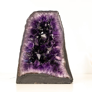 Puede incluir: Una geoda de amatista grande con un exterior gris oscuro y una cavidad llena de cristales de color púrpura intenso. Los cristales están agrupados, creando un efecto brillante. La geoda se encuentra sobre una superficie blanca.