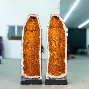 Op de afbeelding: Twee hoge, open geodes met een levendig oranje-gele kristallen binnenkant en een witte rand. De geodes staan op een zwarte basis, tentoongesteld in een galerie.