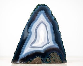 May include: A triangular-shaped agate geode slice with concentric bands of white, light blue, and dark blue. The edges are a dark teal color, and the base has a rough, earthy texture. This decorative stone is ideal for home decor.