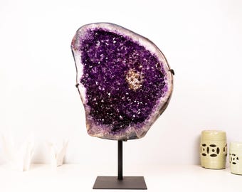 May include: A large amethyst geode with a purple and white crystal formation on a black metal stand. The geode is displayed on a white surface.