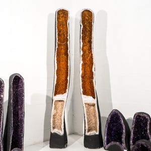 Op de afbeelding: Twee grote, hoge geodekristallen, een met oranje en witte kristallen erin en de andere met paarse kristallen erin. De kristallen staan op een witte achtergrond.