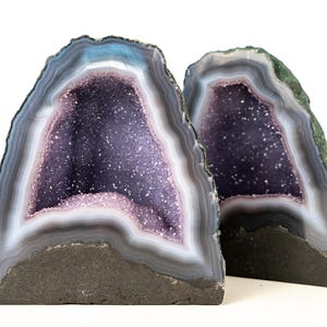 May include: Two amethyst geode bookends. The geodes have a dark purple interior with sparkling crystals. The outer edges are banded with shades of grey, white, and blue. The bases are a dark grey colour.