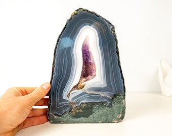 May include: A large, polished agate geode with a striking display of colors. The outer edge is a deep blue-gray, transitioning to white and then a vibrant purple crystal center. The base has a green-gray hue.