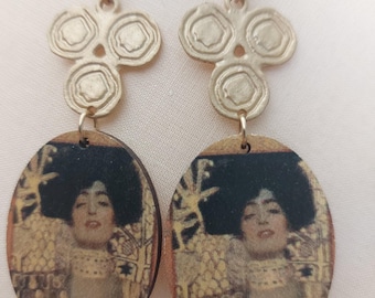 May include: A pair of gold-toned earrings. Each earring features an oval pendant with a portrait of a woman with dark hair and a gold-colored background. The earrings have a decorative top with three circular designs and a hook.