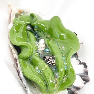 May include: A decorative oyster shell with a vibrant green, abstract glass design. The glass art piece features a flowing, organic shape with embedded beads and a small seashell. The shell's edges are white and black.