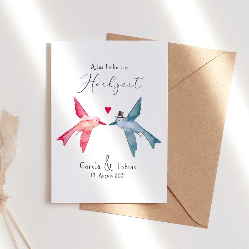 May include: A white card with a watercolor illustration of two hummingbirds, one pink and one blue, facing each other. The text "Alles Liebe zur Hochzeit" is at the top of the card, and the text "Carola & Tobias 14. August 2021" is at the bottom.