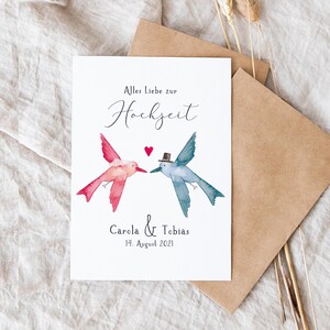 May include: A white card with a watercolor illustration of two hummingbirds facing each other. The hummingbird on the left is pink and the hummingbird on the right is blue. The text "Alles Liebe zur Hochzeit" is written in black cursive at the top of the card. Below the hummingbirds, the text "Carola & Tobias 14. August 2021" is written in black.