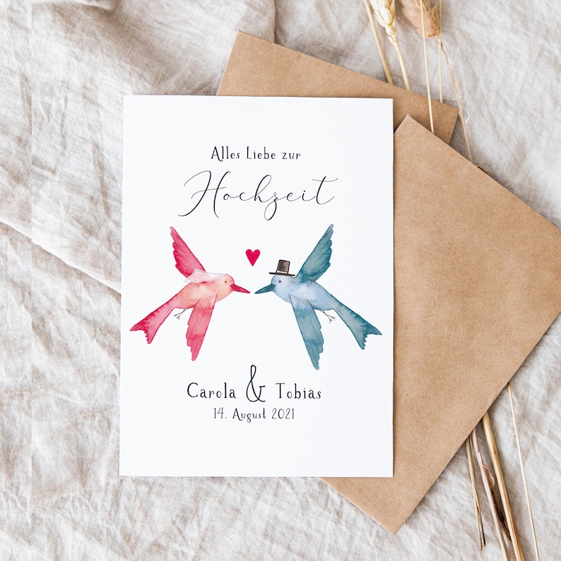May include: A white card with a watercolor illustration of two hummingbirds facing each other. The hummingbird on the left is pink and the hummingbird on the right is blue. The text "Alles Liebe zur Hochzeit" is written in black cursive at the top of the card. Below the hummingbirds, the text "Carola & Tobias 14. August 2021" is written in black.