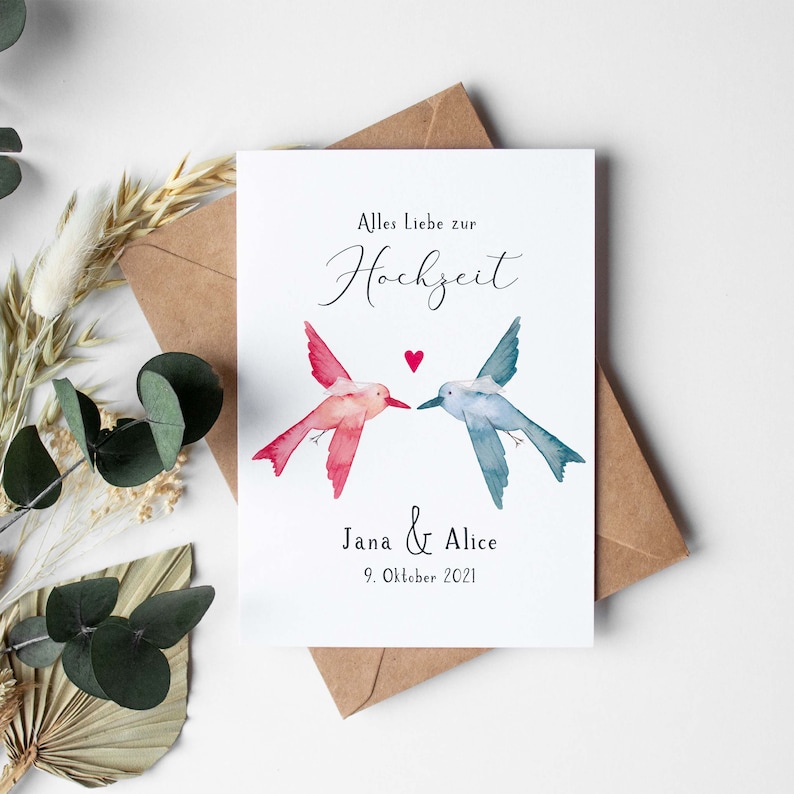 May include: A white card with a watercolor illustration of two hummingbirds facing each other. The hummingbirds are pink and blue. The text on the card reads "Alles Liebe zur Hochzeit" and "Jana & Alice 9. Oktober 2021".