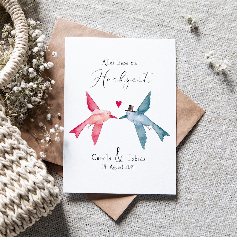 May include: A white card with a watercolor illustration of two hummingbirds, one pink and one blue, with a red heart between them. The text "Alles Liebe zur Hochzeit" is written in black cursive script. Below the birds, the text "Carola & Tobias 14. August 2021" is written in black.