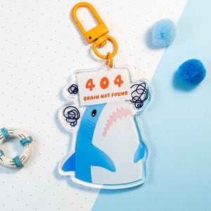 Puede incluir: Un llavero acrílico transparente con un tiburón de dibujos animados con el texto "404 BRAIN NOT FOUND". El tiburón es azul y blanco con una expresión preocupada. El llavero tiene un cierre naranja. También, un pequeño salvavidas y dos pompones azules.